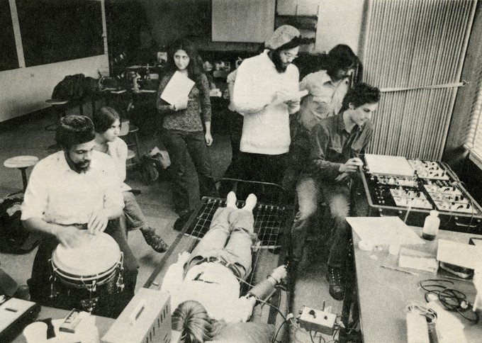 Black and white photograph of a laboratory with a class. A figure lies on a cot in the center, a wire attached to their arm. A bearded figure in a hat plays a hand drum while seated on the left. In the background, several students look on, holding papers and taking notes. One of them is seated on the right in front of a panel of dials and switches.