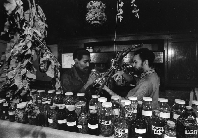 Black and white photograph of two men surrounded by containers of herbs.