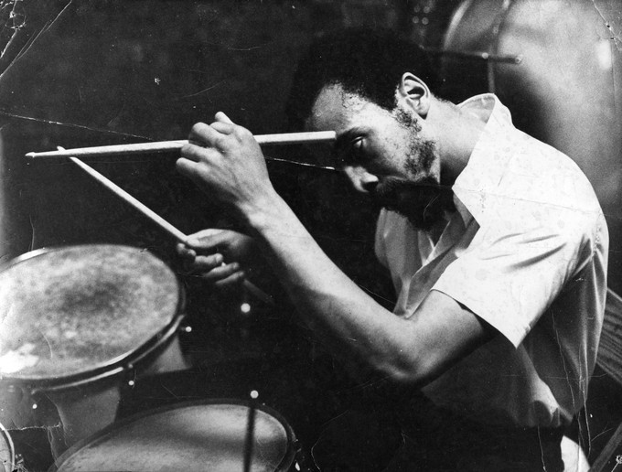 A black and white image of a person sitting at a drum set. He is holding up two drumsticks, and staring intently.
