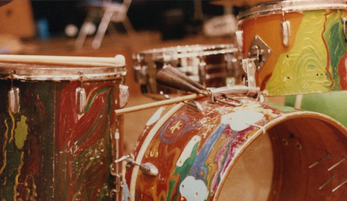 A close-up view of a hand-painted percussion or drum set. The  drums are painted in red, orange, white, and green hues. Drum sticks sit atop two of the drum heads while a third drum head positioned in the center of the picture frame has its membrane open. 