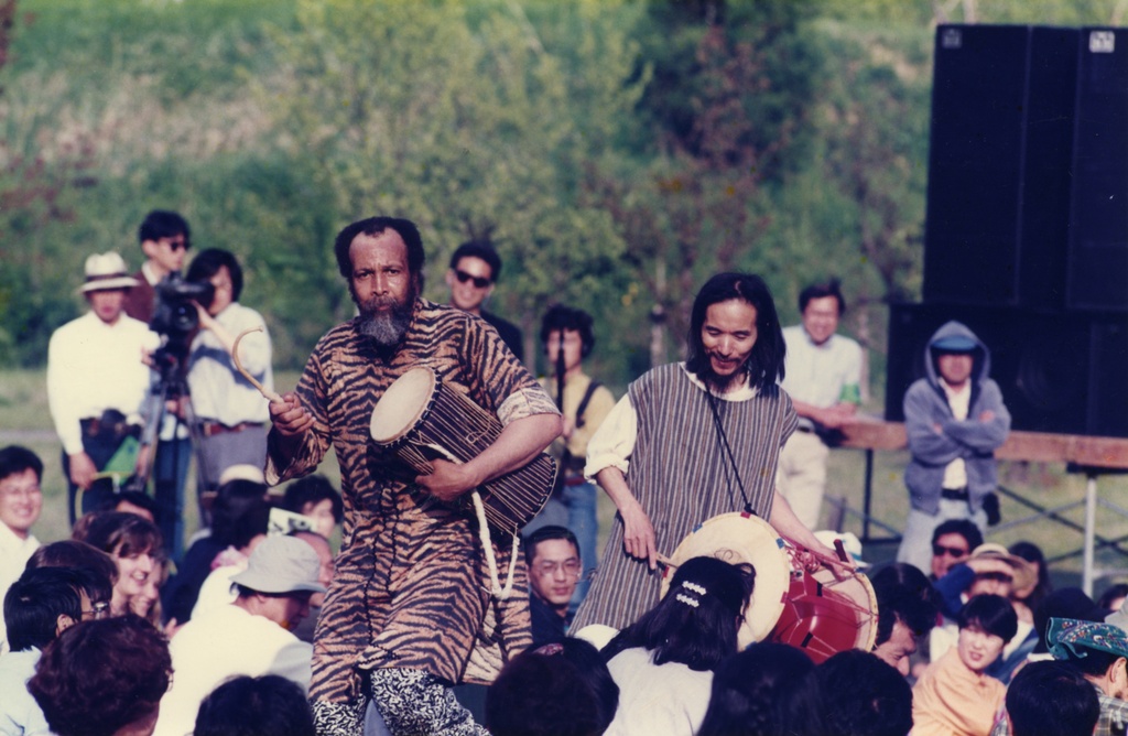 Color photograph of two figures holding drums amidst a crowd in an outdoor performance. The figure on the left is wearing a tiger print tunic, the figure on the right wears a striped tunic.&nbsp;
