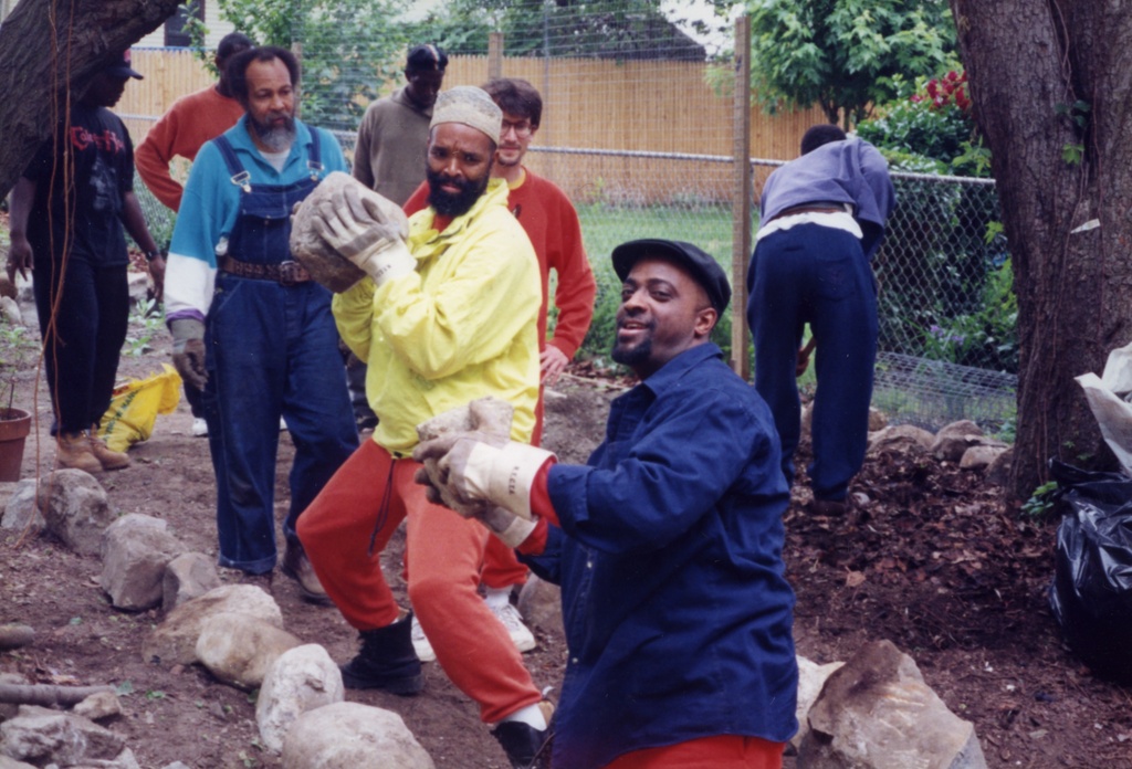 Color photograph of several figures wearing blue, yellow, and red clothing standing in a backyard between a pathway of stones. The two figures closest to the camera carry medium sized grey stones.&nbsp;