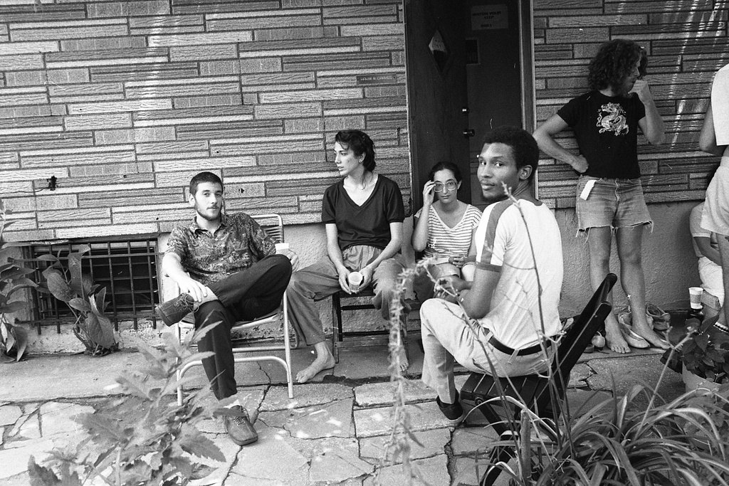 A black and white photograph of a group of people sitting and standing out on the patio of a home. Several plants are visible in the foreground, and a figure on the right in a white t-shirt twists around to look at the camera.&nbsp;
