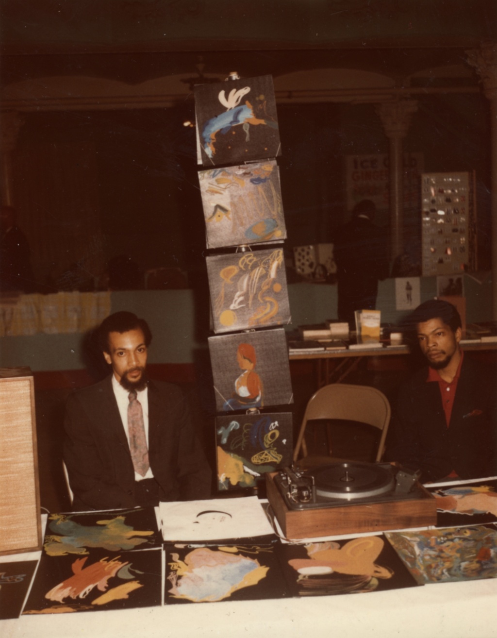 Photograph of two men sitting behind a table surrounded by hand painted album covers.