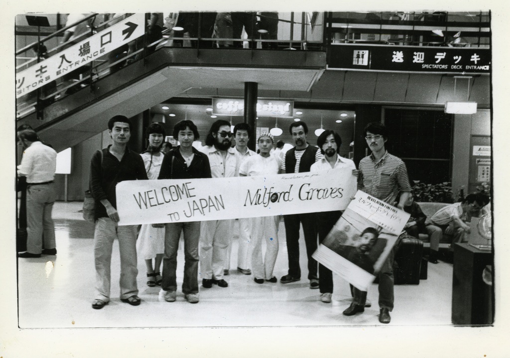 <p>A black and white photograph of a group of figures standing in an airport. They hold a long rectangular sign which reads "Welcome to Japan Milford Graves." A figure on the right holds another poster featuring an image of a person. Japanese characters are visible on signage in the background.&nbsp;</p>