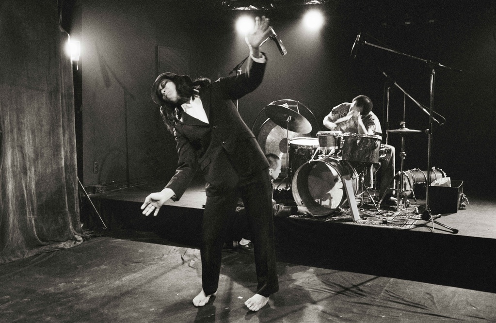 Black and white photograph of a performance. In the foreground, a barefoot figure in a suit leans to their right side. A drummer is visible in the background.