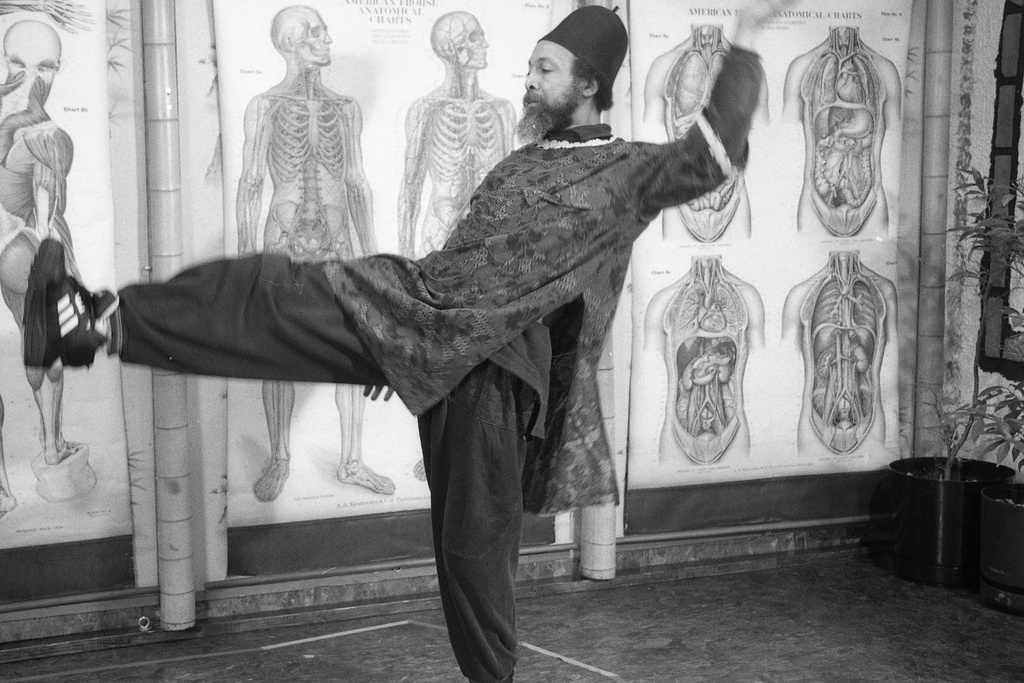 A black and white photograph of a bearded figure in a cylindrical hat and long tunic. They balance on their left leg, raising their right leg up to hip height while also raising their left hand up in the air. In the background anatomical drawings of human figures are visible.&nbsp;