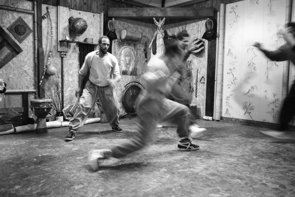 A black and white photograph of several figures lunging. The figure in the center moves so quickly that their image is blurry. In the background, several drums, a gong, and a poster of a figure in a feathered headress are visible.&nbsp;