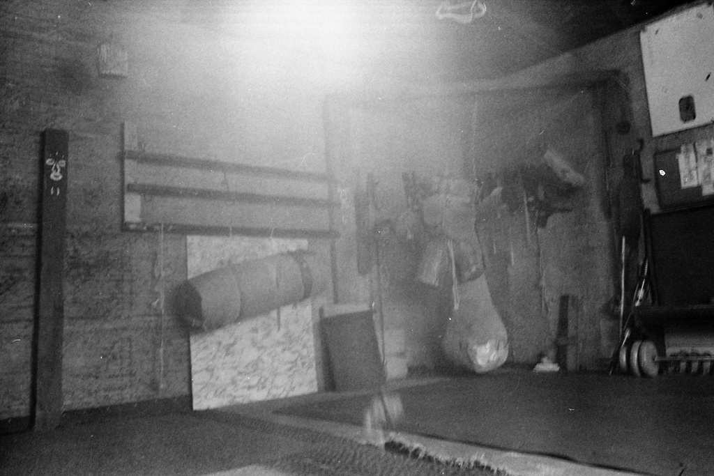 A black and white photograph of an interior. The floor is lined with mats, and several suspended punching bags are visible.&nbsp;