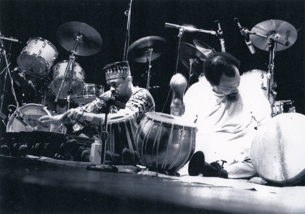 A black and white image of two men kneeling on the ground playing African drums.