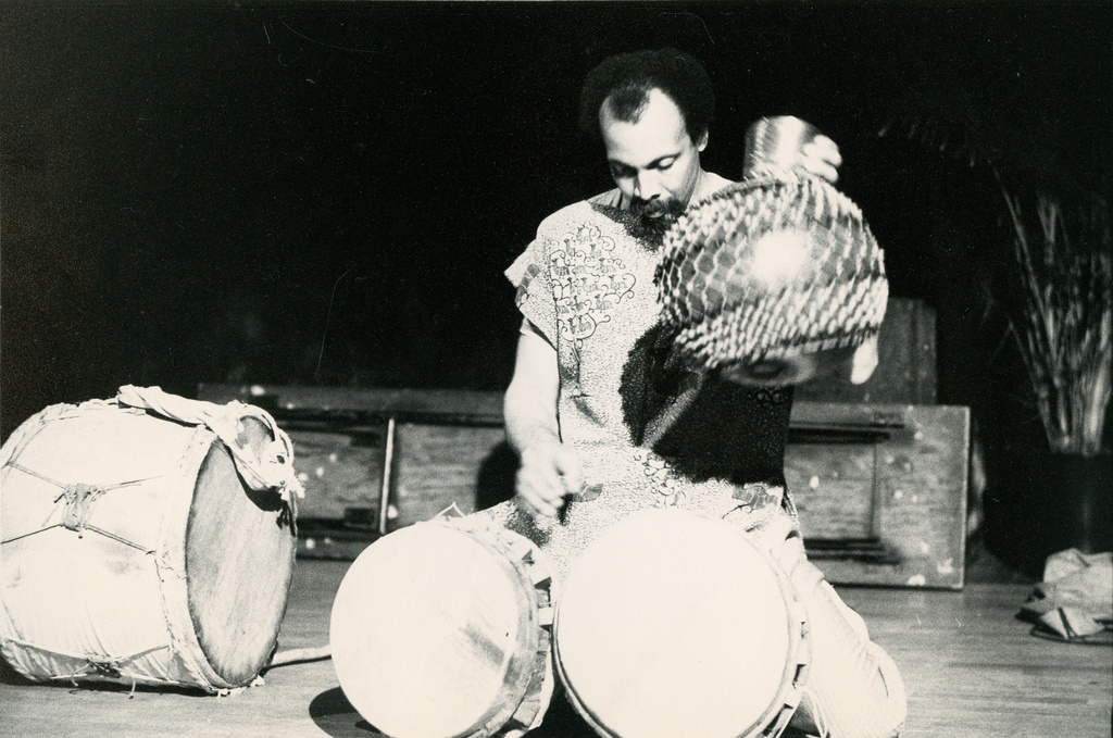 A black and white image of a man sitting on his knees with two African drums placed in front of him. His head looks downward while his left-hand hovers above the drums. His right hand firmly holds the side of a shaker instrument.