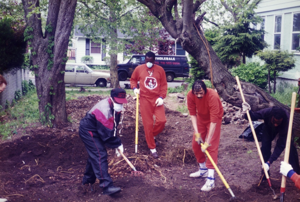 Color photograph of four figures with rakes and hoes standing outside on a pile of dirt. They are framed by a pair of trees.&nbsp;