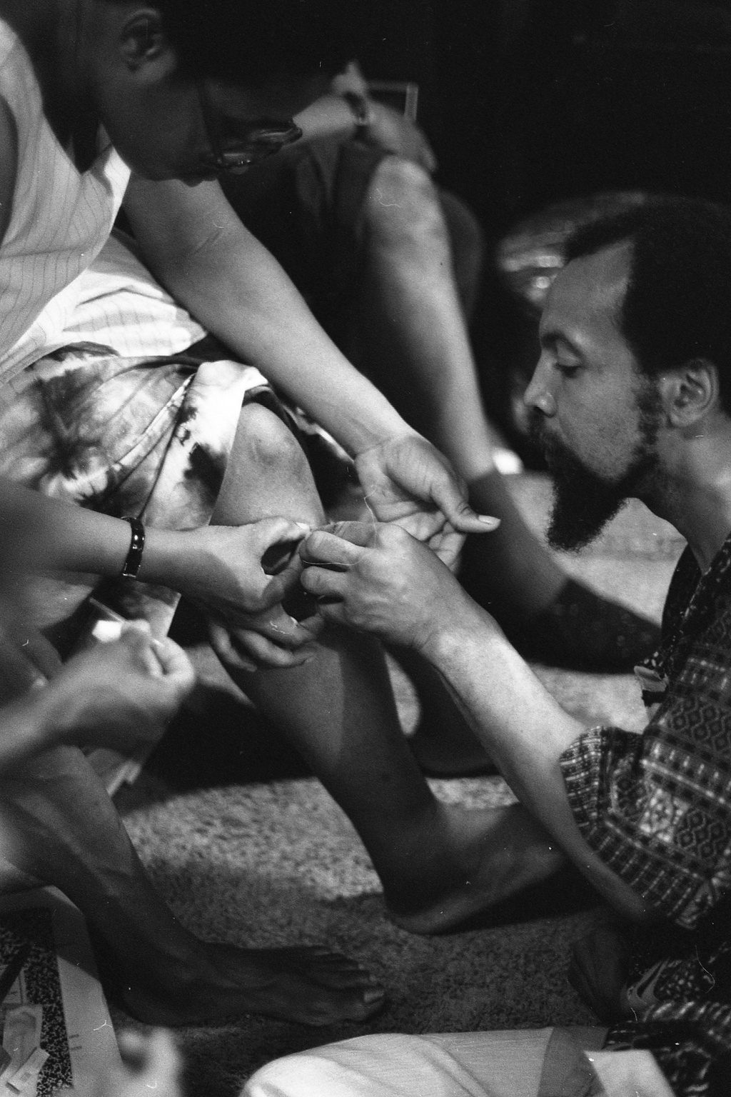 A black and white photograph of a person in a short-sleeved collared shirt with a patchwork pattern performing acupuncture on another person's knee. Both figures are sitting.&nbsp;