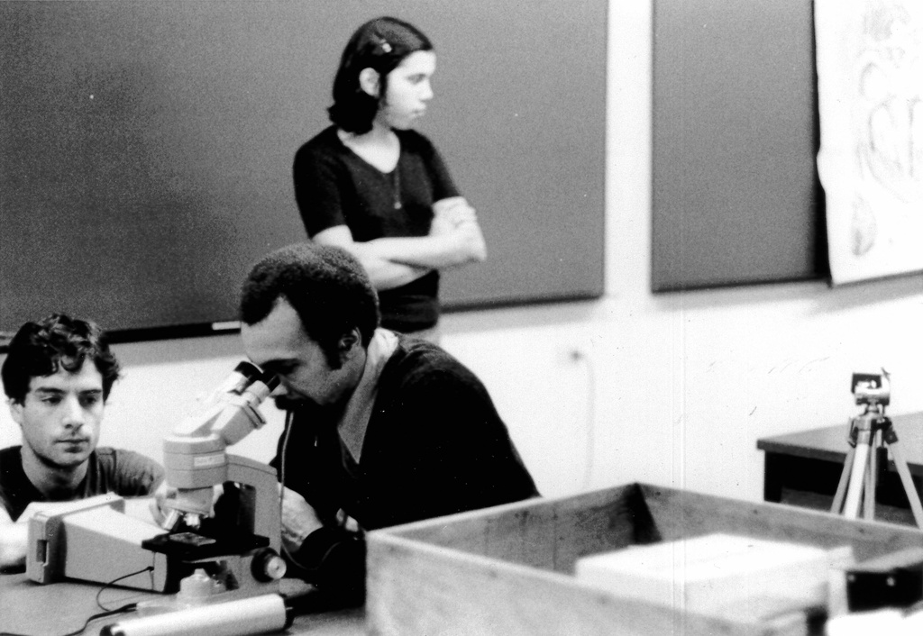 A black and white image depicts an individual studying a subject under a microscope in a classroom environment. Two individuals surround him, one sitting to his left watching intently, and the other standing and facing her head in the opposite direction.&nbsp;